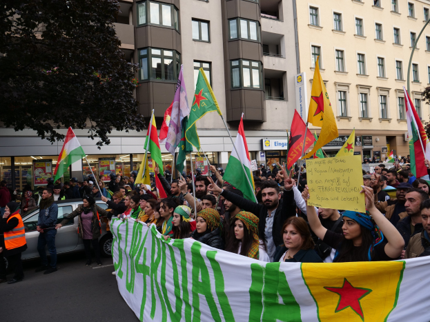 Große Gruppe von Menschen marschiert auf einer Straße mit Fahnen und Spruchbändern, mit einem geparkten Auto auf der rechten Seite und einem Baum auf der linken Seite, vor Gebäuden mit sichtbaren Fenstern und Beschriftungen.