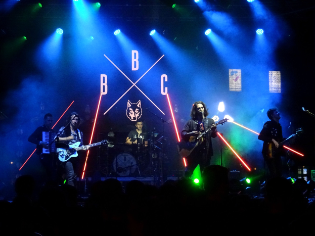 Eine Band performt auf der Bühne mit Gitarristen, die in Mikrofone singen, ein Schlagzeuger im Hintergrund und ein beleuchtetes Publikum darunter.