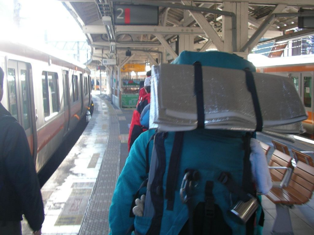 Ein Bahnhofsgebäude mit Menschen neben einem Zug auf der linken Seite, einem Stand in der Mitte, einem weiteren Zug auf der rechten Seite und einer Bank in der rechten unteren Ecke.