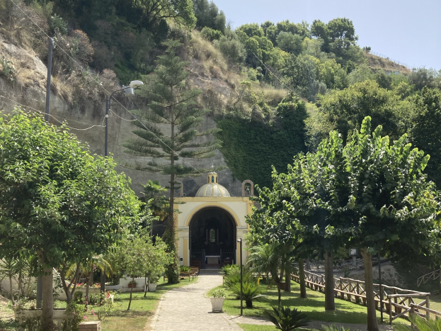 Eine kleine Kirche ist inmitten einer grünen Hügellandschaft eingebettet, umgeben von Bäumen, Pflanzen, Gras, einem Pfad, einem Zaun, Straßenlaternen, Strommasten, Stromkabeln, Hügeln und einem klaren blauen Himmel.
