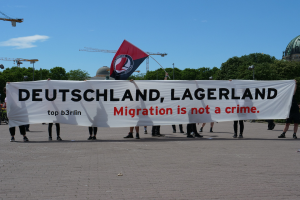 Gruppe von Menschen mit einem Banner 'Deutschland, Lagerland Migration ist kein Verbrechen' und einer Flagge, mit Straßenlaternen, Lichtern, Bäumen, Kränen, einem Gebäude und einem bewölkten Himmel im Hintergrund.