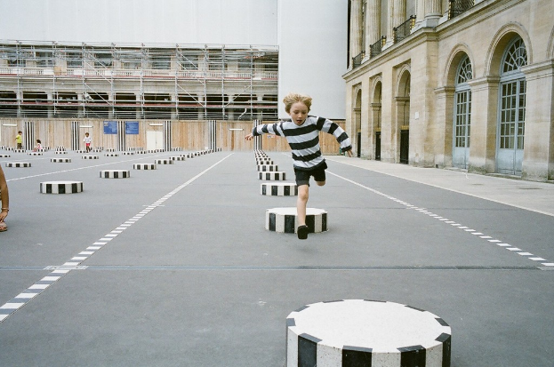 Vier Kinder befinden sich in einem Schulspielplatzbereich mit schwarzen und weißen Betonstufen; ein Kind läuft auf dem Boden.
