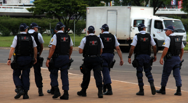 Eine Gruppe von Beamten in Uniform, einschließlich Mützen, Hemden, Schutzwesten, Uhren, Hosen und Schuhen, die auf einer Straße mit einem rechts geparkten Lastwagen in der Nähe eines Zauns gehen. Im Hintergrund sind Gebäude, Bäume, Pflanzen, Pfosten, Banner und eine Brücke oben rechts zu sehen.