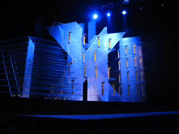 Eine Bühne für eine Preisverleihung mit dunklem Hintergrund, beleuchtet von ein paar Lichtern und mehreren Gegenständen, die mit dem Ereignis in Verbindung stehen.