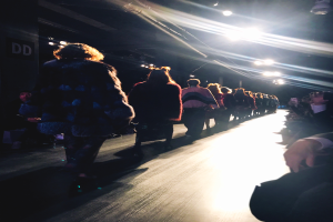 Eine Gruppe von Menschen läuft über einen hell erleuchteten Laufsteg bei einer Fashion Show, während Zuschauer auf beiden Seiten sitzen.