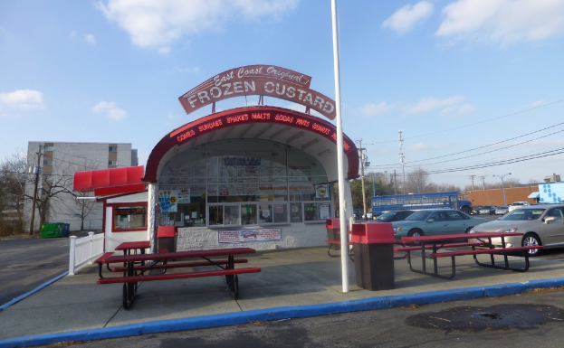 Eine Szene mit Autos, Pfählen, Gebäuden, Bänken, einem Schuppen mit einer Tafel und Plakaten am Schuppen.