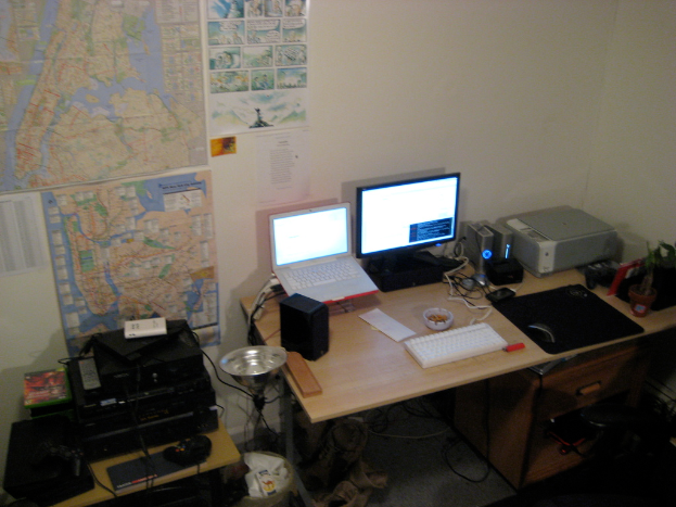 Ein Raum mit einem Tisch, auf dem ein Laptop, Lautsprecher, Monitor, Tastatur, Ball, Maus, Scanner und Papiere liegen, und einem weiteren Tisch mit Elektronikgeräten im Hintergrund, mit Diagrammen und Postern an der Wand dahinter.