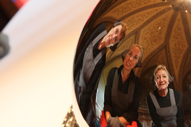 Ein reflektierender Ball auf einem wei├čem Tisch zeigt das Spiegelbild von drei lachenden Frauen, die sich leicht b├╝gen, mit einem Dach im Hintergrund.