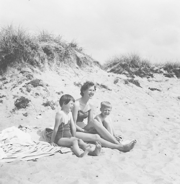 Schwarz-weißes Foto einer Frau und zweier Kinder auf einer Stranddecke sitzend, mit Sand, Gras und Himmel im Hintergrund.