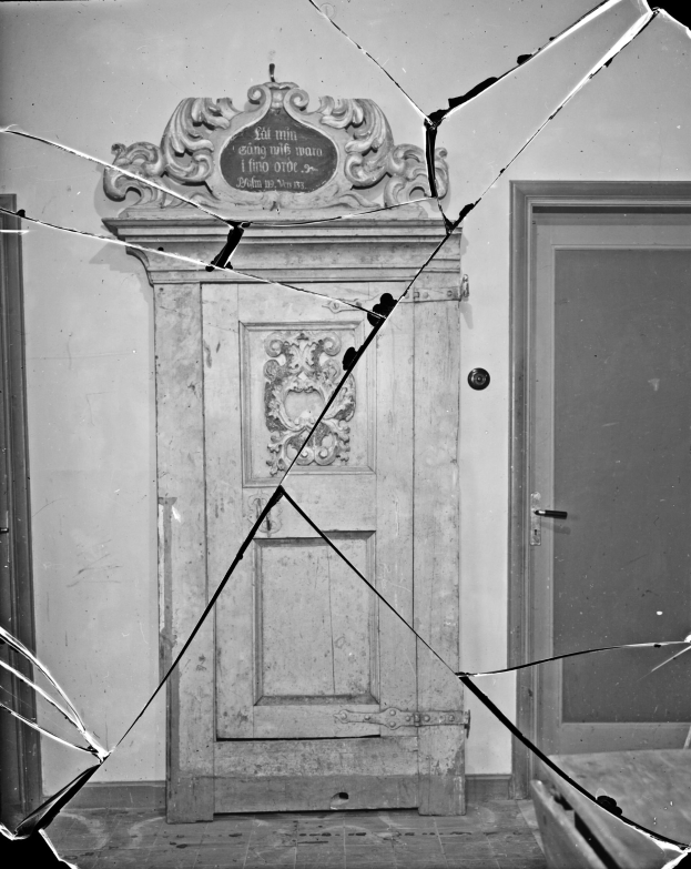 Ein Schwarz-Weiß-Foto einer zerbrochenen Tür in einem Raum, mit zerbrochenem Glas drumherum und einer Wand im Hintergrund.