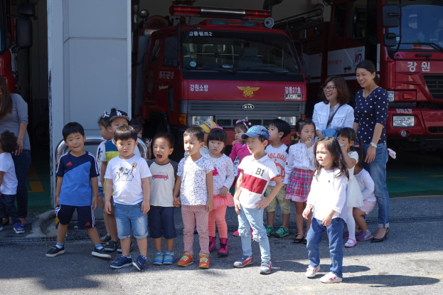 Eine Gruppe von Kindern vor mehreren Feuerwehrautos auf einer Feuerwache, einige tragen Mützen.