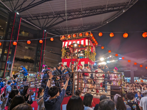 Eine Menschenmenge auf einer beleuchteten Bühne mit Laternen, einige halten Kameras, umgeben von einem Geländer, mit Bäumen und einem blauen Himmel im Hintergrund bei Nacht, möglicherweise während eines japanischen Festivals.