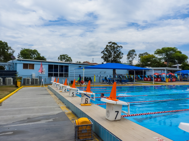 Großes Freibad mit mehreren Schwimmern, Schwimmbahnen, Verkehrskegeln, Liegestühlen, Sonnenschirmen, einem Gebäude mit Fenstern, einer Flagge, Bäumen und einem bewölkten Himmel.