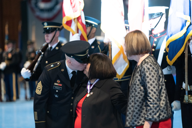 Ein Paar umarmt und küßt sich, während eine Gruppe uniformierter Personen mit Mützen im Vordergrund steht, einige halten Fahnen.