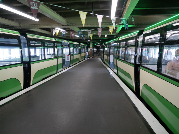 Grüner und weißer Zug fährt auf Schienen neben einem Bahnsteig mit sichtbaren Passagieren im Inneren, unter einer Decke mit Lampen und dekorativen Fahnen an einem Bahnhof in München, Deutschland.