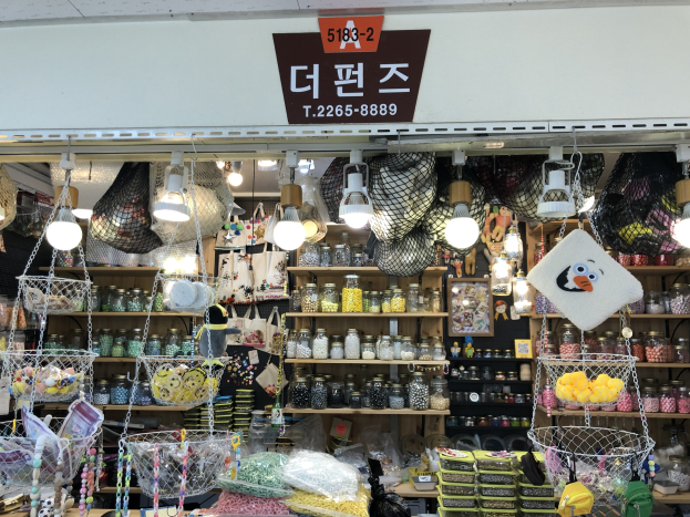 Ein unordentlicher Laden in Seoul zeigt verschiedene Gegenstände wie Körbe, Lampen, Flaschen und Spielzeuge auf Regalen, mit einer Tafel oben, die anzeigt, dass es ein top-bewerteter koreanischer Shop ist.