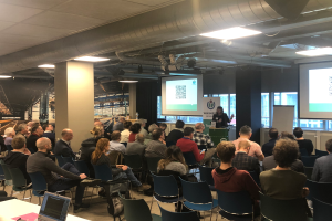 Eine große Gruppe von Menschen sitzt auf Stühlen vor einem Projektionsschirm, ein Redner steht neben einem Mikrofon und einem Gerät auf einem Ständer in einem Raum mit einer Tafel, Säulen, Fenstern, Deckenleuchten und einer Decke.