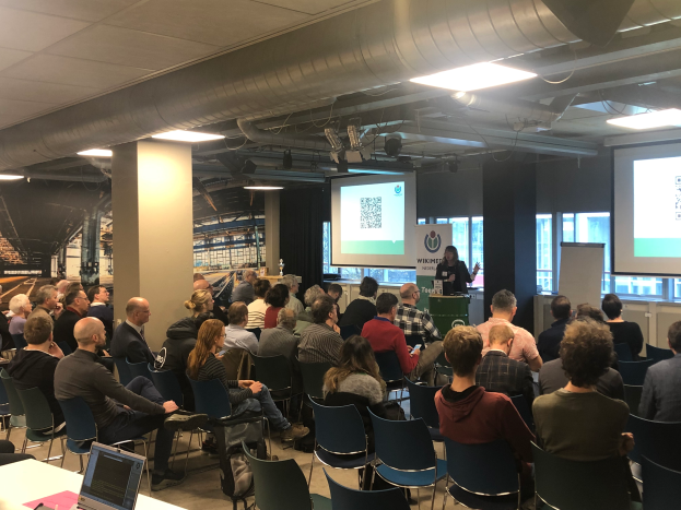 Eine große Gruppe von Menschen sitzt auf Stühlen vor einem Projektionsschirm, ein Redner steht neben einem Mikrofon und einem Gerät auf einem Ständer in einem Raum mit einer Tafel, Säulen, Fenstern, Deckenleuchten und einer Decke.