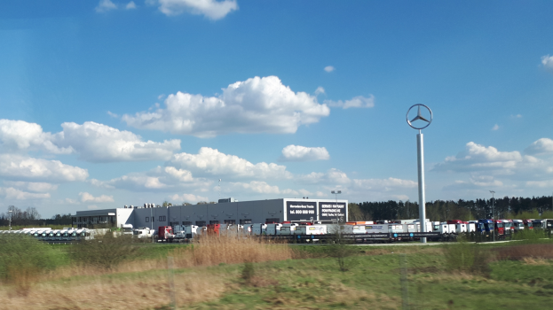 Ein Mercedes-Benz-Werk in Deutschland mit Fahrzeugen auf der Straße, umgeben von Gras, Pflanzen, Bäumen, Gebäuden und Polen, von denen einer ein Logo hat, unter einem bewölkten Himmel.