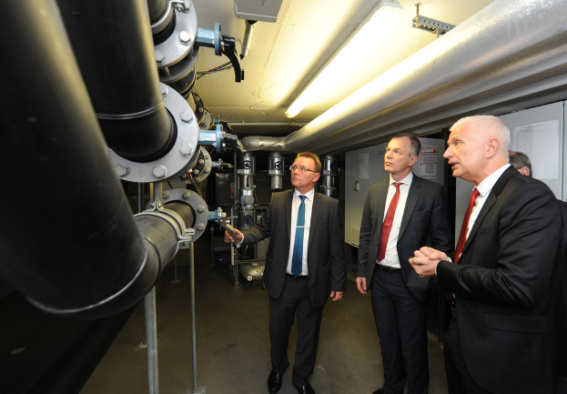 Vier Personen rechts, drei mit Mänteln, mit großen Rohrleitungen links.