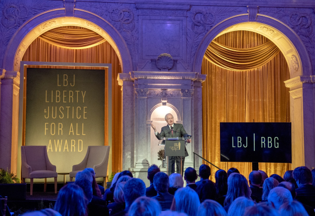 Ein Mann steht an einem Rednerpult mit einem Mikrofon und spricht vor einem Publikum, das auf Stühlen sitzt, in einem Raum mit Pflanzen, Vorhängen und einer Tafel mit Text dahinter.