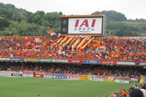 Ein Fußballspiel wird in einem Stadion mit einer großen Zuschauermenge, grünem Rasen, einem Torpfosten, Bannern, Fahnen, einem großen Bildschirm, Bäumen und einem klaren blauen Himmel gespielt.