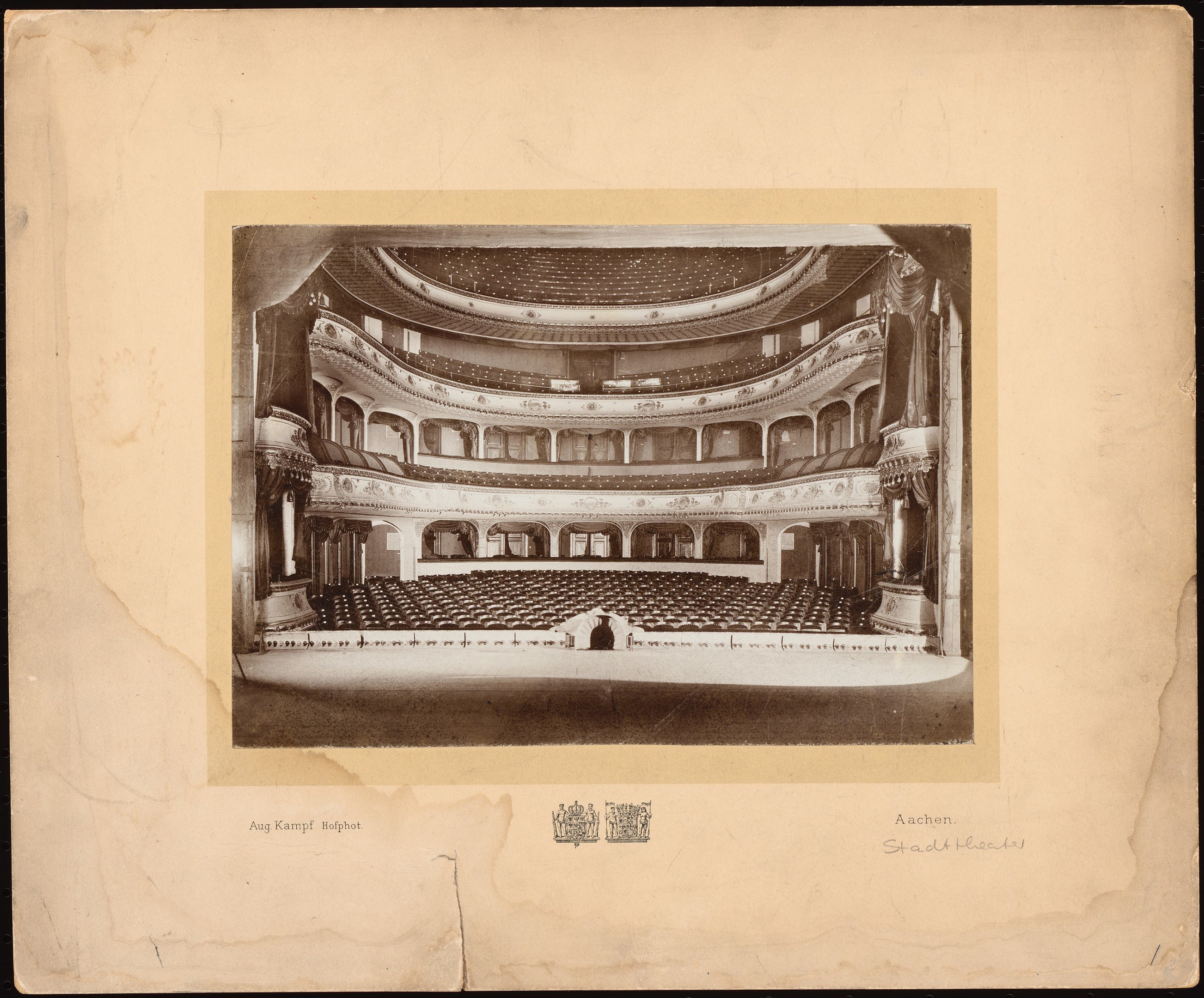 Altes Schwarz-Weiß-Foto des Auditoriums der Royal Albert Hall, das die Bühne, Sitzreihen, Säulen, Geländer und Text/Logo unten zeigt.