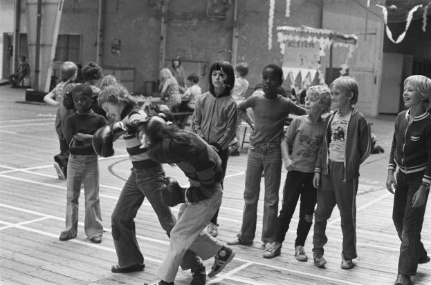 Eine Gruppe von Kindern, die auf einem Basketballfeld spielen, mit zwei kämpfenden Personen in der Mitte und einigen Personen auf den Tribünen im Hintergrund, dargestellt in Schwarz-Weiß.