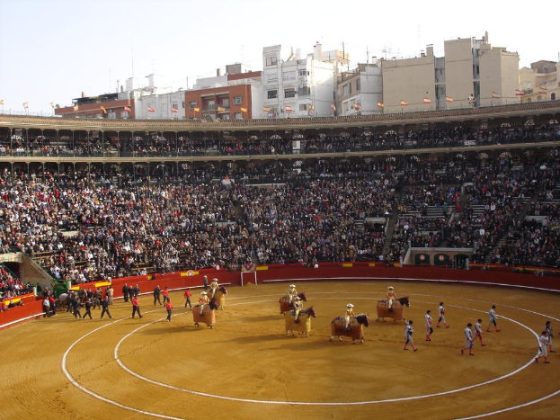 Menschen gehen auf dem Boden, einige reiten auf Pferden, andere sitzen im Stadion und schauen zu, Fahnen und Gebäude sind im Hintergrund unter dem Himmel zu sehen.