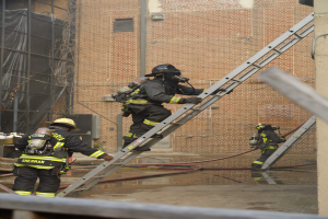 Eine Gruppe von Feuerwehrleuten in Helmen und Zylindern klettert an einer Leiter vor einem Backsteingebäude hoch, mit Rohren und einem Metallstab auf dem Boden und einem Gebäude mit Fenstern und einem Netz im Hintergrund.