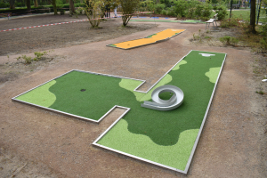 Minigolfplatz in einem Park mit Grünfläche, Menschen in der Nähe und Gebäuden im Hintergrund bei klarem Himmel.