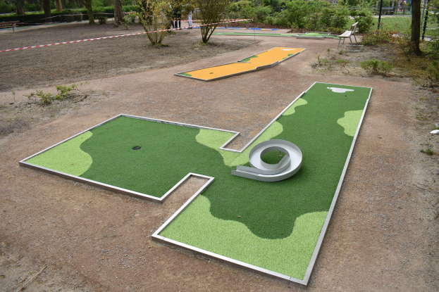 Minigolfplatz in einem Park mit Grünfläche, Menschen in der Nähe und Gebäuden im Hintergrund bei klarem Himmel.