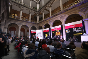 Eine Gruppe von Menschen, die auf einer Reihe von Stühlen vor einem Publikum sitzen, wobei einige Individuals mit Kameras ausgestattet sind, während einer Pressekonferenz für das CDMX 2017-Event, das ein Podium mit einem Mikrofon in der Mitte, Banner mit Text und Bildern, Säulen, Geländer, Fenster und eine Wand im Hintergrund zeigt.