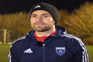 Ein Mann in einer blauen Jacke und Mütze steht auf einem Fußballfeld während eines Trainingsspiels, mit Bäumen und einem Tor im Hintergrund.