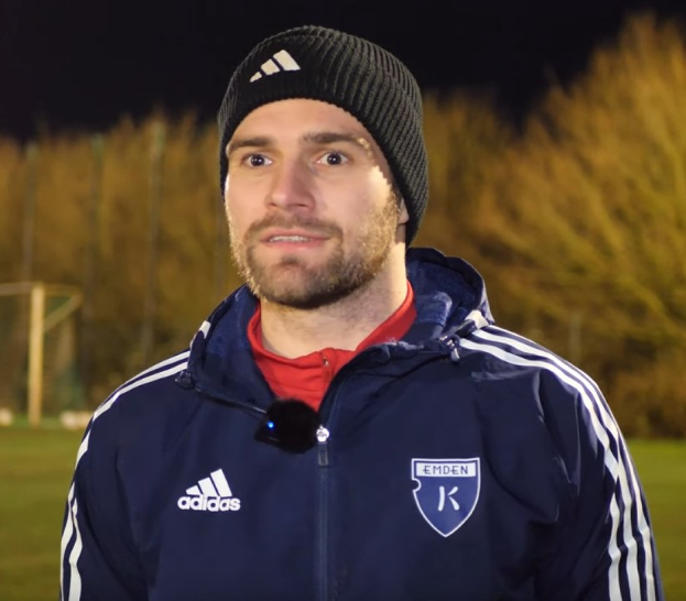 Ein Mann in einer blauen Jacke und Mütze steht auf einem Fußballfeld während eines Trainingsspiels, mit Bäumen und einem Tor im Hintergrund.
