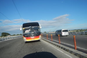Ein Shuttlebus fährt auf einer Autobahn neben einem Gewässer entlang, mit Geländern auf beiden Seiten, Bäumen, Türmen und einem bewölkten Himmel im Hintergrund.
