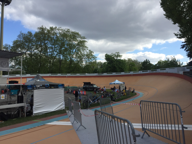 Gruppe von Radfahrern auf einer Schotterstrecke bei den UCI BMX-Weltmeisterschaften 2018, mit Zelten, Fahrzeugen, Geländern, Bäumen und einem bewölkten Himmel im Hintergrund.