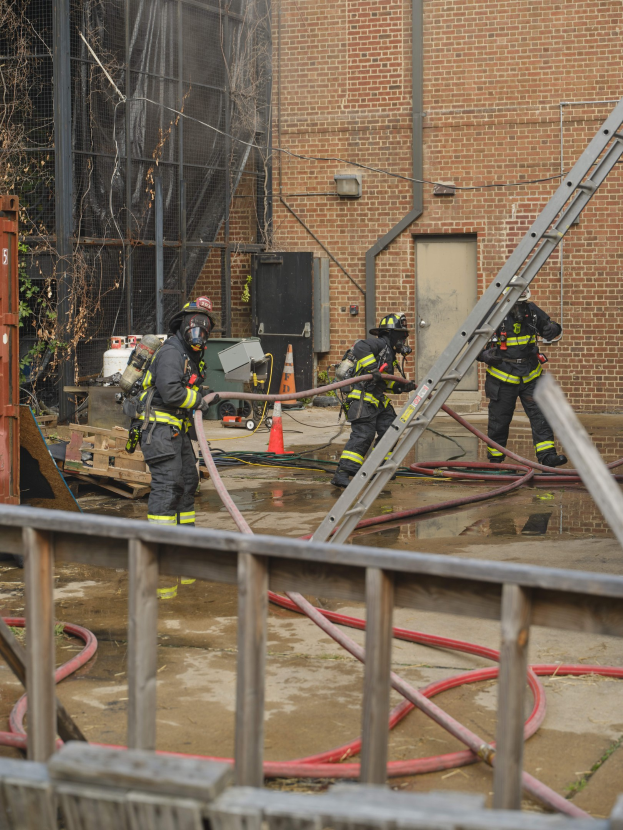 Feuerwehrleute mit Helmen arbeiten daran, ein Gebäude Feuer zu löschen, während sie Schläuche halten, mit einem Metallzaun, Rohren, einem Container, einer Verkehrskegel, Gegenständen, Fenstern, Tür, Metallrahmen, Drähte, Baum und Himmel sichtbar.