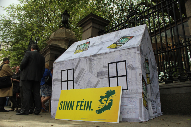 Ein Zelt und ein Werbeplakat sind auf dem Boden platziert, mit einer Menge daneben, einem Eisenzaun, Säulen, Straßenlaternen, Bäumen und Himmel im Hintergrund.