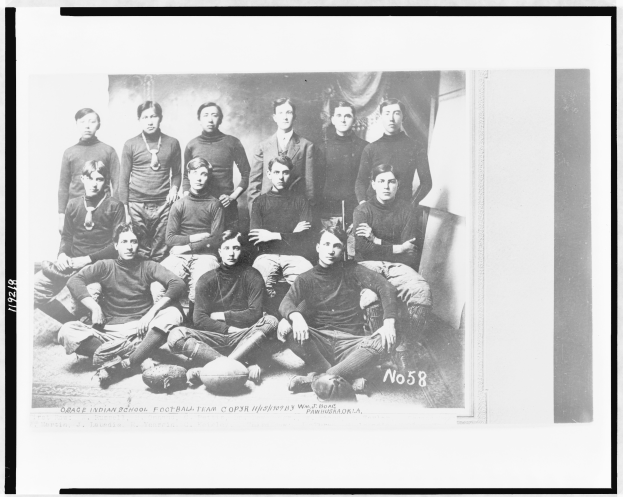 Ein Schwarz-Weiß-Foto einer Fußballmannschaft, mit einigen Spielern sitzend und einigen stehend, Bälle auf dem Boden verstreut und Text unten.