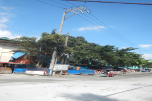Eine Stadtstraße mit fahrenden Fahrzeugen, Fußgängern auf dem Gehweg, Gebäuden, Bäumen und einem bewölkten Himmel.