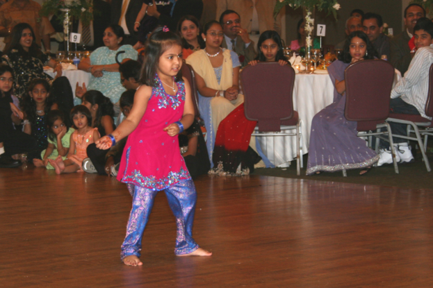 Ein Mädchen tanzt im Vordergrund auf einem Holzboden, während im Hintergrund viele Menschen an Tischen sitzen, die mit weißen Tischdecken und kleinen Pflanzen bedeckt sind.