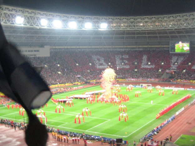 Ein Stadion mit Flutlicht, Eisengittern, Zuschauern, stehenden Personen, Werbetafeln, Anzeigetafeln, einer Überwachungskamera und verschiedenen Dekorationen einschließlich Flaggen.