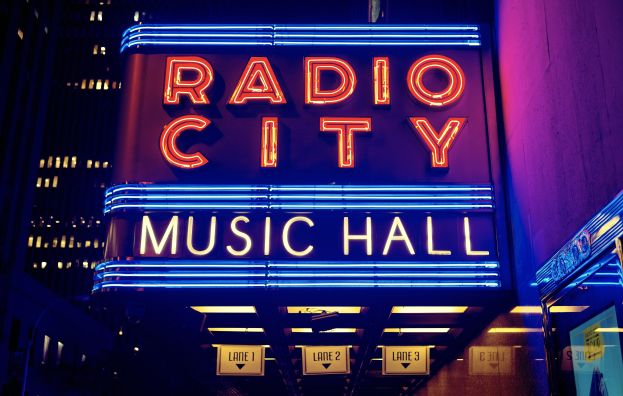 Hell erleuchtetes Radio City Music Hall in New York City mit hohen umliegenden Gebäuden und einem "Radio City"-Schild auf der rechten Seite.