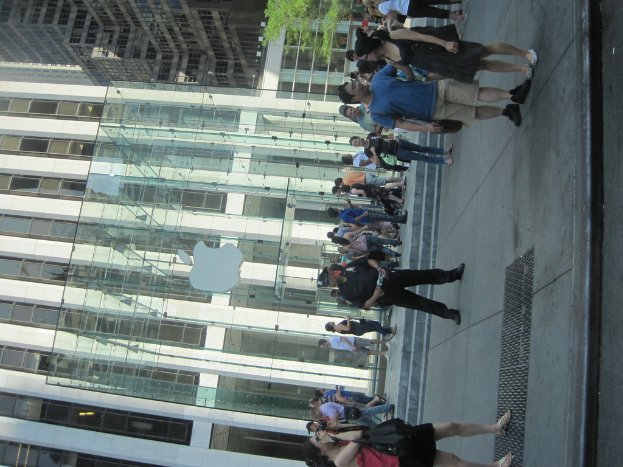 Ein modernes Gebäude mit Glaswänden und Säulen, das ein Logo zeigt, mit vielen Menschen davor und einer Treppe, die hinaufführt, während einige Menschen auf dem Gehweg rechts stehen und Zweige mit Blättern oben im Bild zu sehen sind.