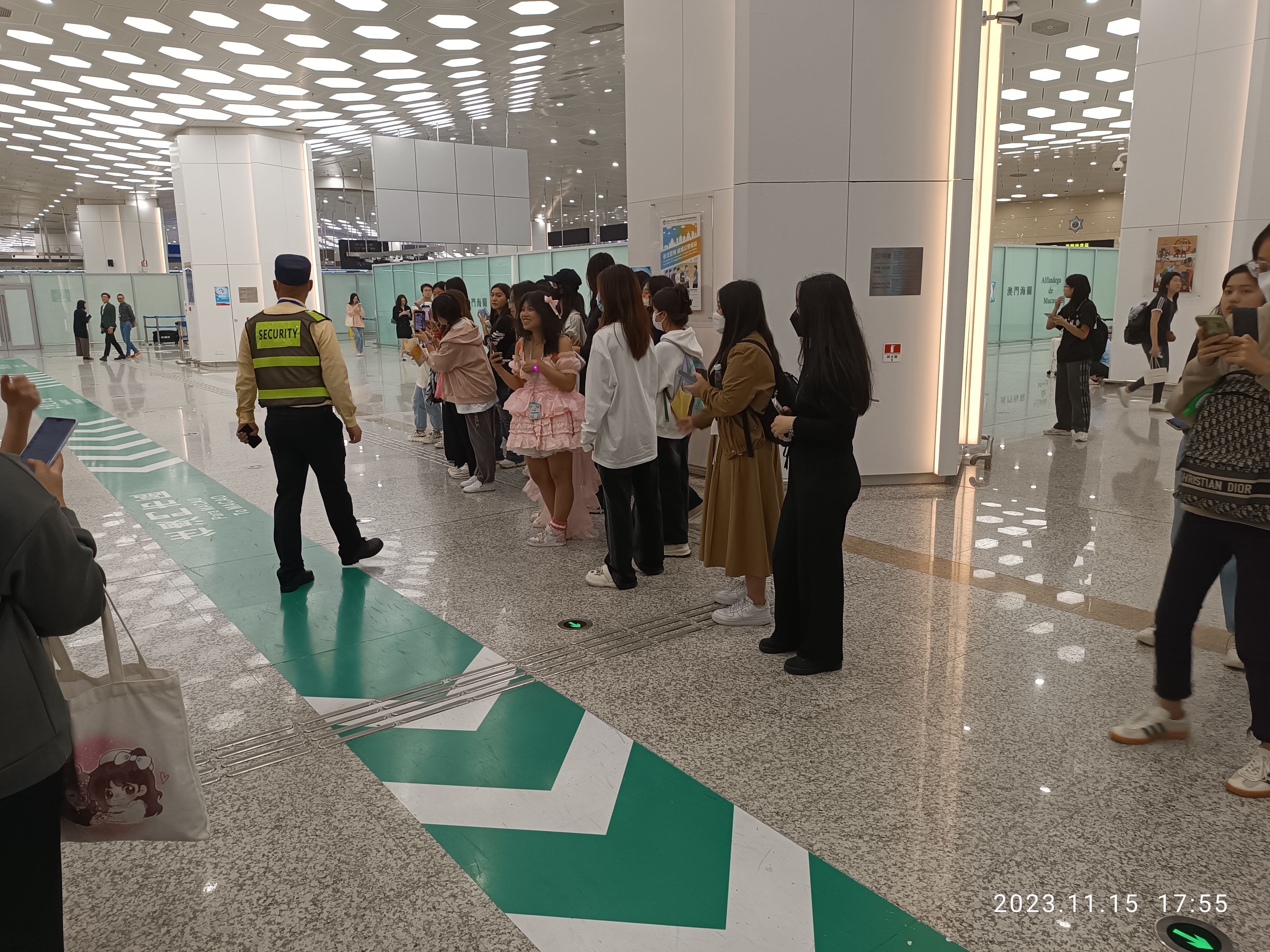 Eine Gruppe von Menschen steht in einer Reihe an einem Flughafen-Sicherheitscheck-in, einige halten Taschen und Mobiltelefone, mit Säulen im Hintergrund und Lampen an der Decke und einem Wasserzeichen am unteren Rand des Bildes.