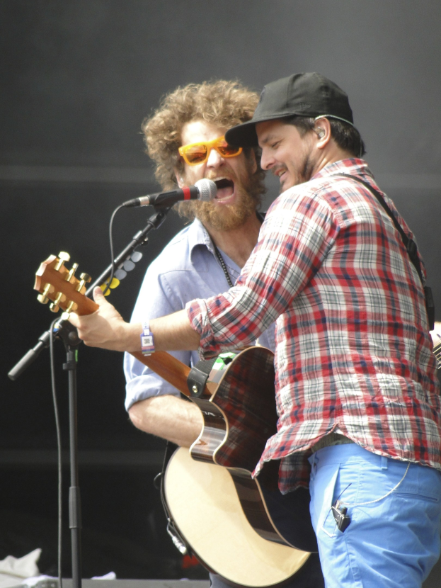 Zwei Männer auf der Bühne, einer in rot-blau kariertem Hemd und blauen Hosen, der andere mit Schutzbrille und schwarzer Mütze, beide singend in Mikrofone, während sie Gitarren spielen.