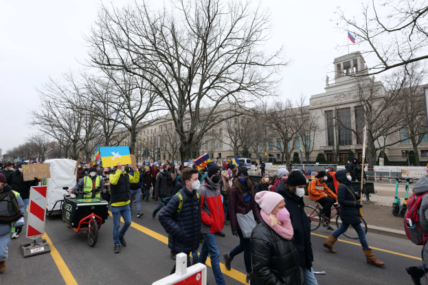 Eine große Gruppe von Menschen nimmt an einer Protestmarsch auf einer Straße in Washington, D.C. teil, wobei einige Schilder und Banner halten, andere Fahrräder fahren und Schilder, Bäume und ein klarer blauer Himmel im Hintergrund zu sehen sind.