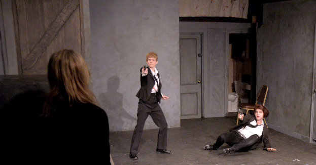 Zwei Frauen in schwarzen Anz√ľgen, eine sitzend auf dem Boden eine Waffe haltend, die andere stehend und eine Waffe haltend, mit einer dritten Frau in der N√§he, in einem Raum mit einem Stuhl, einer Toilette und einer hölzernen Tür.