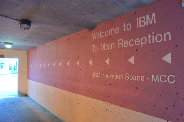 Eine Wand mit dem Text "Willkommen bei IBM" beleuchtet von Deckenlampen, mit einer Tür im Hintergrund beschriftet mit "IBM Innovation Space MCC".